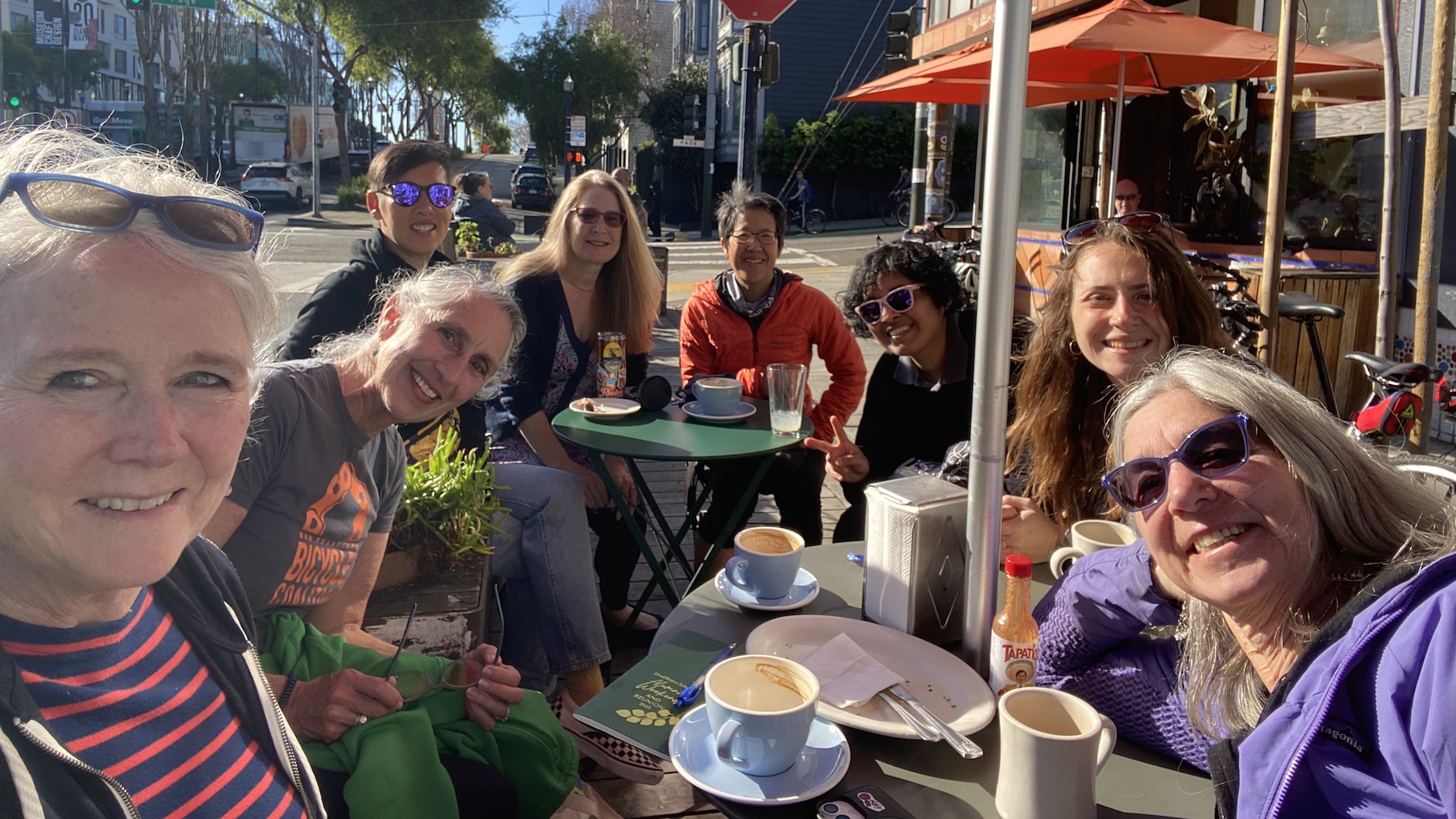 9 people gathered around patio tables in a city.