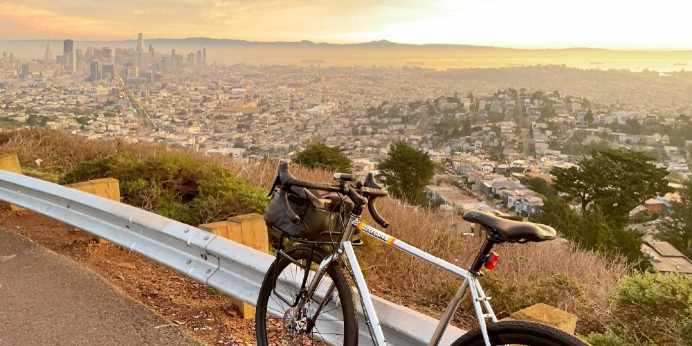 A silver bike with front rack and bag leaning against a guardrail at Twin Peaks, overlooking an early morning view of San Francisco