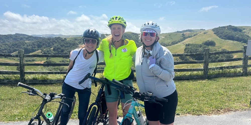 three people in helmets stand in front of a wood fence and rolling green hills