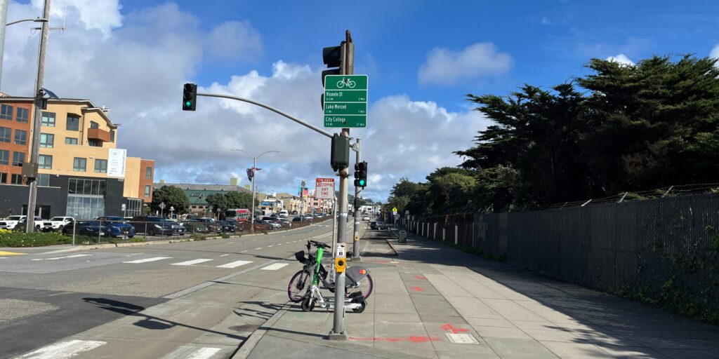 photo of Sloat Blvd with green sign showing bicycle directions to nearby destinations. A bike share bike and lime scooter are next to the sign.