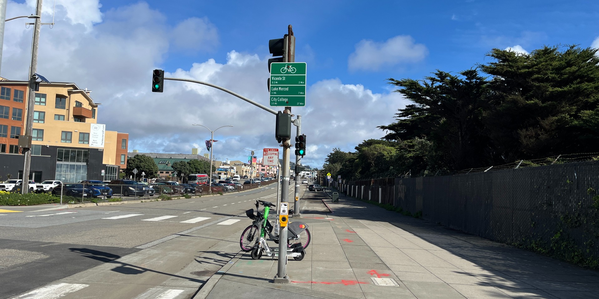 photo of Sloat Blvd with green sign showing bicycle directions to nearby destinations. A bike share bike and lime scooter are next to the sign.