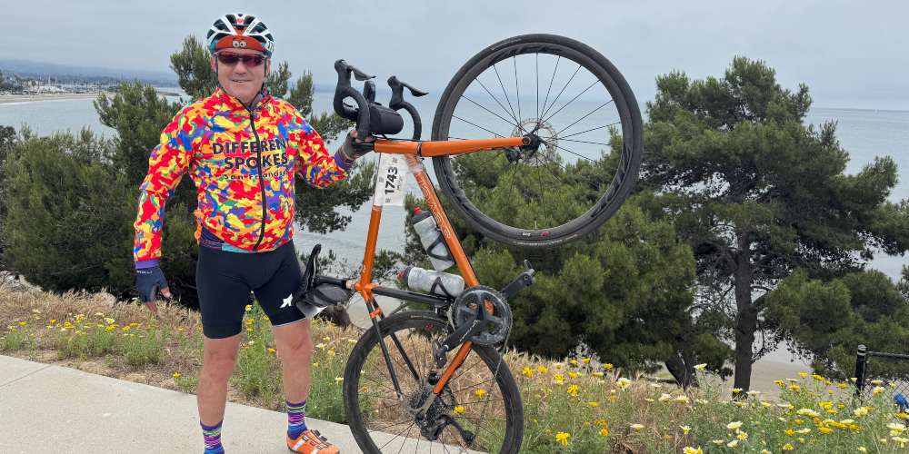 A person in a colorful bike kit stands holding their bike. He is in front of trees and water