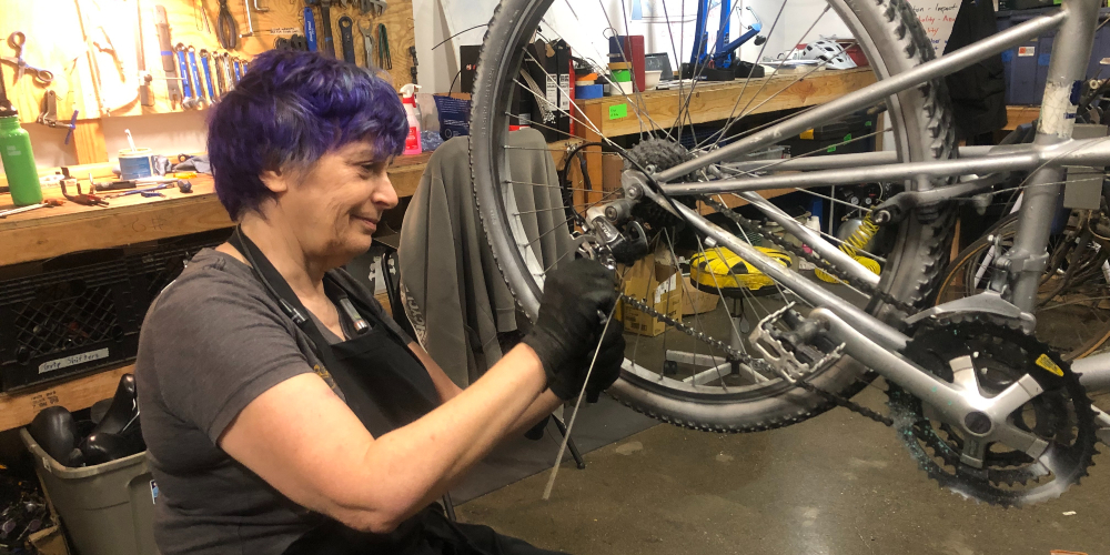 A person with purplish blue hair works on the back tire of a silver bike