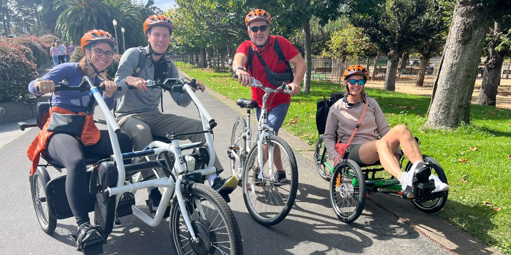 Four people smiling in a park, three of them are on adaptive bikes and one is on a standard bike.