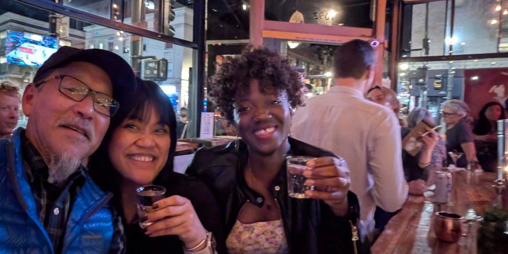 three people in a bar smiling, two of them are holding up drinks