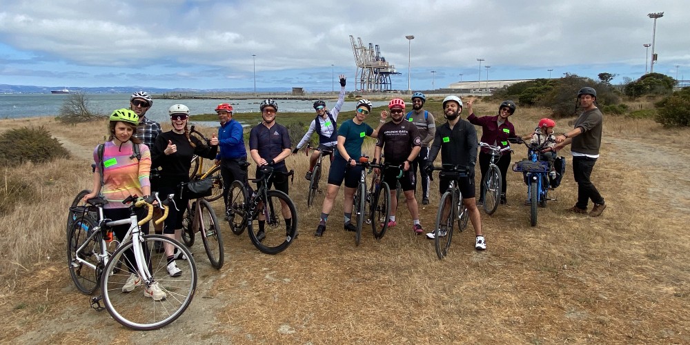 A crowd of people with bikes stand in grass in front of the bay.