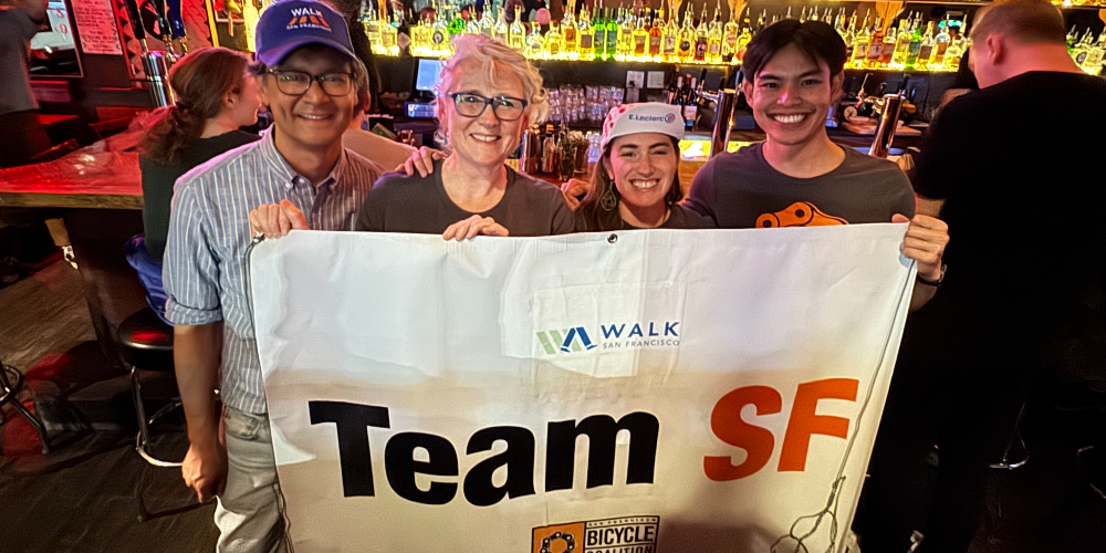 Four people stand in front of a bar, they're holding a banner that reads "Team SF"