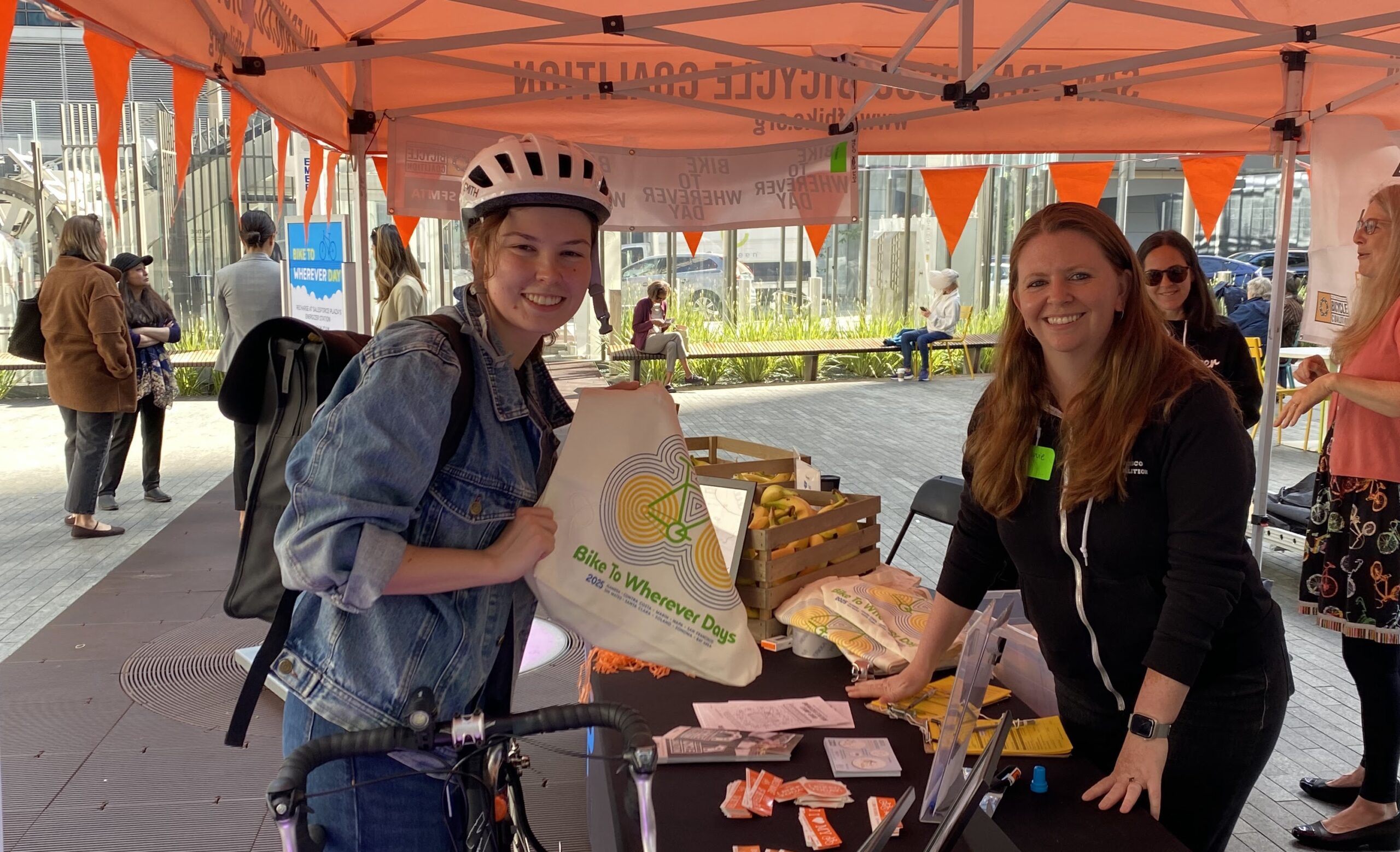 two people smiling, they're separated by a table. one is holding a tote bag with a bike on it