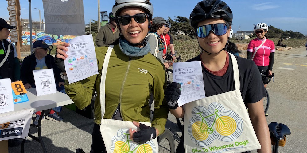 two people wearing helmets and sunglasses posing holding up tote bags with a bike design and papers that read "BTWD energizer station stamp card"