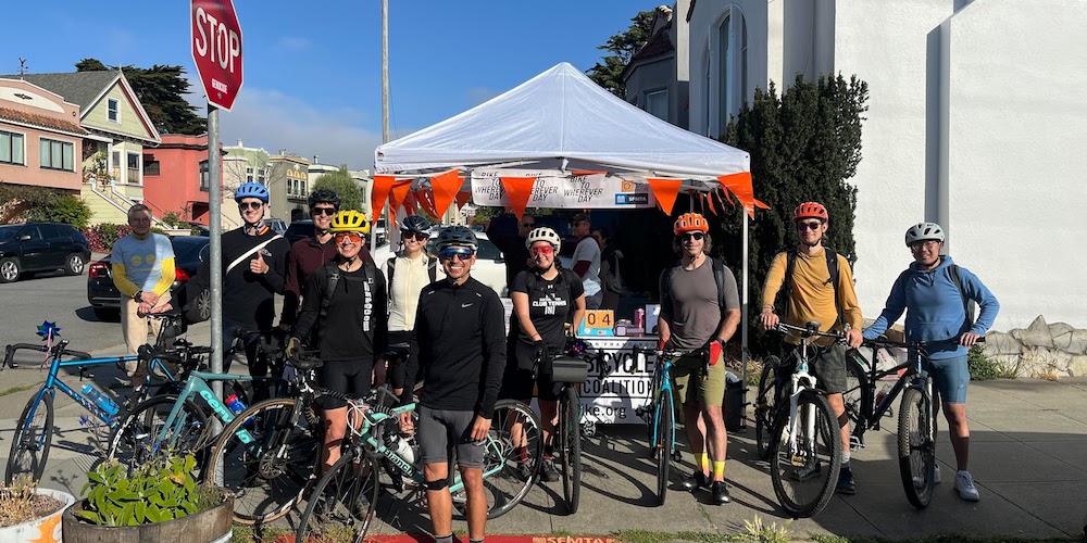 A group of people with bikes stand in front of a tent with orange flags decorating it