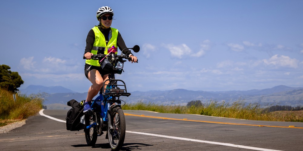 a person on an e-bike, wearing a high visibility vest, rides on a road in front of hills in the distance