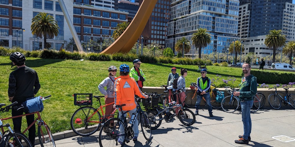 a group of people with bikes stand in front of a lawn with a large sculpture of a bow and arrow