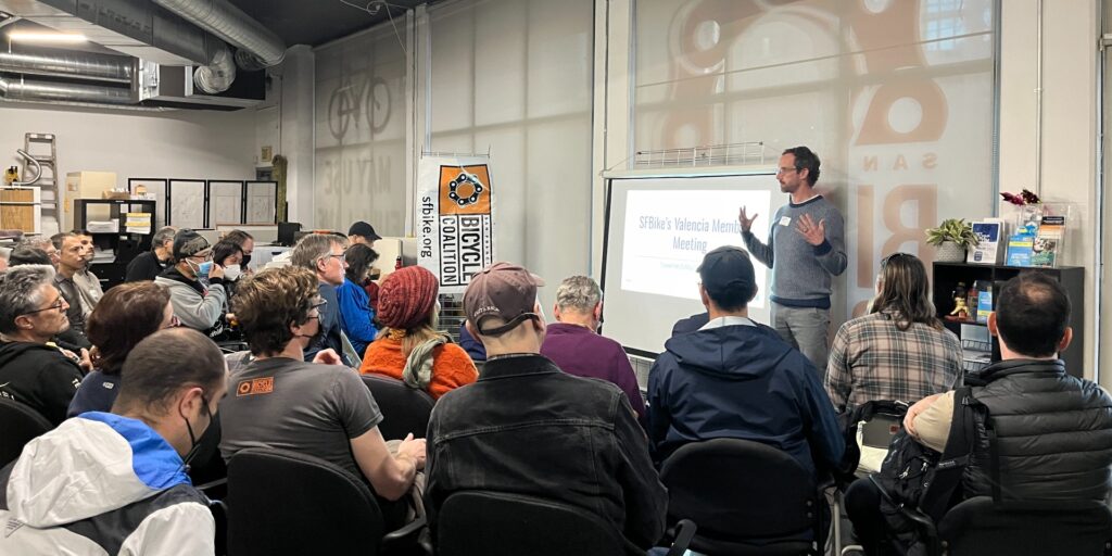 Image of large group sitting in chairs looking at a projector screen and a speaker