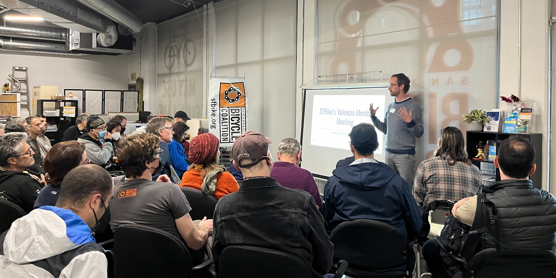 Image of large group sitting in chairs looking at a projector screen and a speaker