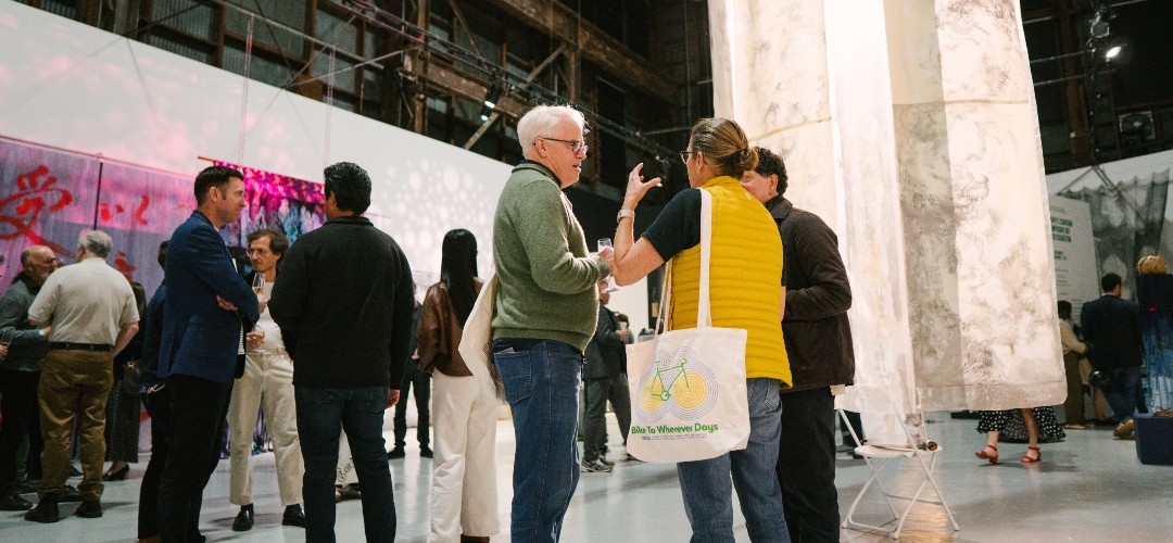 A group of people, one with a tote bag with a bike graphic, standing together in a gallery and chatting
