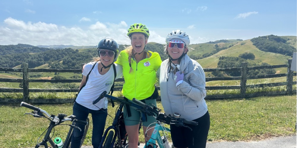 Three people with their bikes stand in front of a wooden fence and rolling green hills