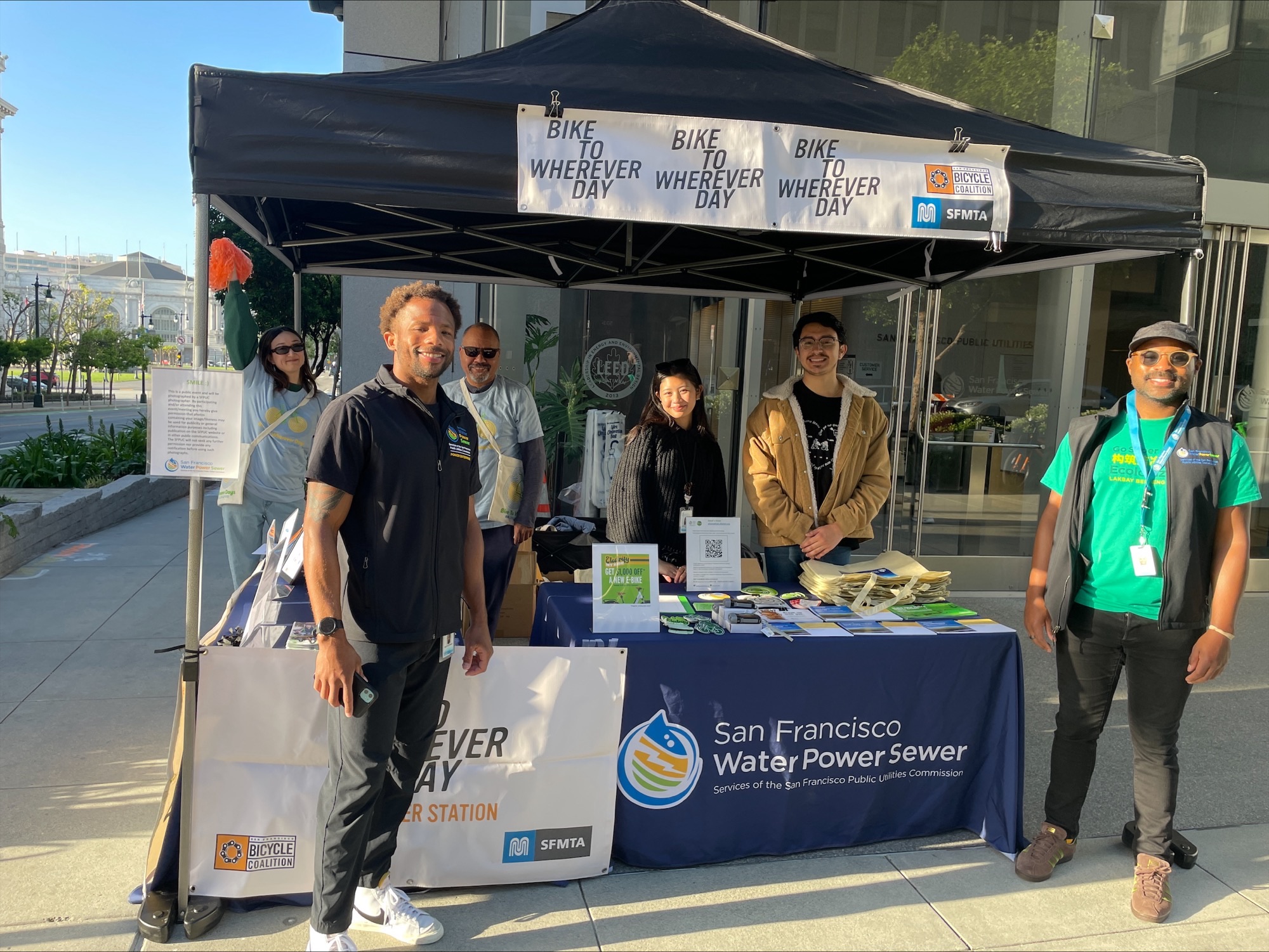 Bike WD 3 (2) A group of people standing together in a tent. There's a banner on the tent that reads "Bike to Wherever Day Energizer Station"
